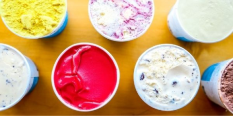 a bird's-eye view of seven open pint containers of brightly colored ice cream on a light wooden surface